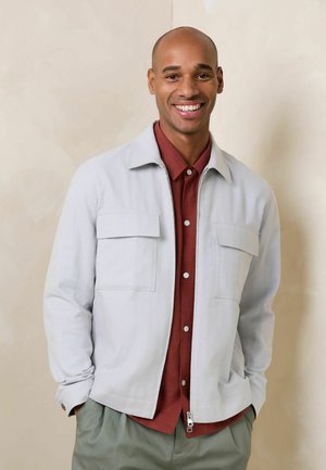 Lächelnder glatzköpfiger Mann trägt hellgraue Reißverschlussjacke über einem rostfarbenen Hemd mit Knöpfen und graue Hose, steht mit Händen in den Taschen.