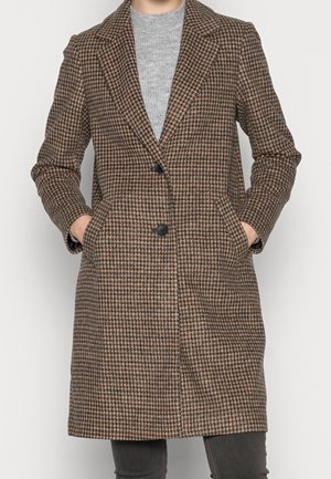 Lang bruin en zwart jas met houndstooth-patroon, revers, twee zwarte knopen en zijzakken, gedragen over een grijze trui en donkere broek.
