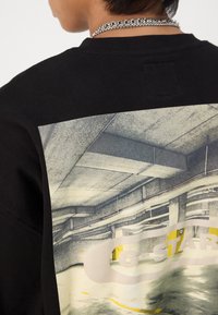 Μαύρη μπλούζα με κουκούλα που έχει μια μεγάλη εκτύπωση στην πλάτη απεικονίζοντας σκηνή από χώρο στάθμευσης με κείμενο "G-Star". Μείγμα βαμβακιού, κανονική εφαρμογή.