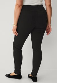 Sorte, strækbare leggings med høj talje, to baglommer og en slim fit, parret med sorte flade sko. Glat tekstur.