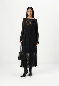 Μαύρο δαντελένιο φόρεμα με μακριά μανίκια, λουλουδάτο μοτίβο, διαφανή υφή και σφιχτή μέση. Συνδυασμένο με μαύρα μποτάκια με τακούνι και μικρή τσάντα.