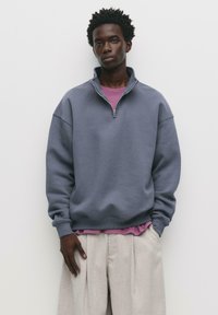 Felpa grigia in fleece con colletto a zip parziale, vestibilità rilassata e polsini a coste. Indossata sopra una maglietta rosa chiaro a strati e pantaloni beige.