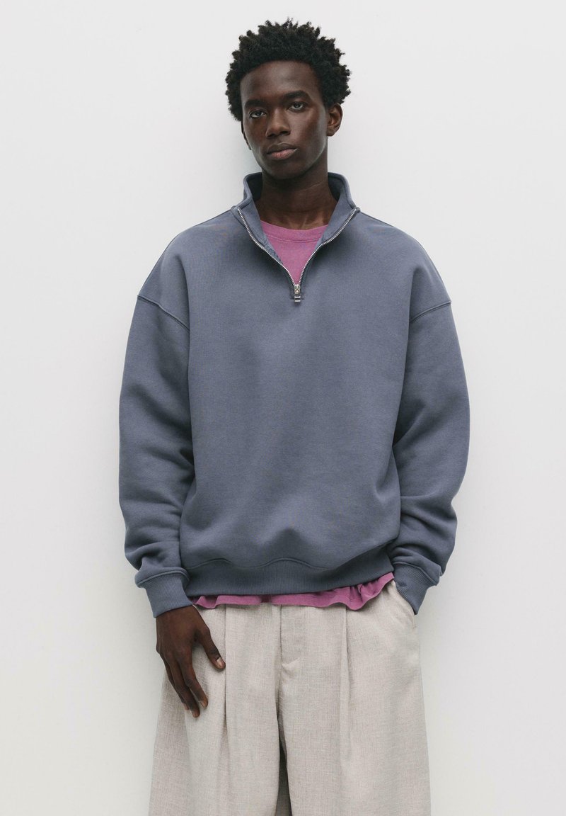 Felpa grigia in fleece con colletto a zip parziale, vestibilità rilassata e polsini a coste. Indossata sopra una maglietta rosa chiaro a strati e pantaloni beige.