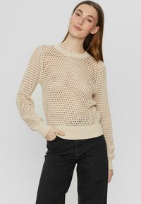 Beige kötött pulóver, laza szövésű anyagból, kerek nyakkivágással, bordás mandzsettákkal és derékrésszel. Sötét farmernadrággal párosítva.