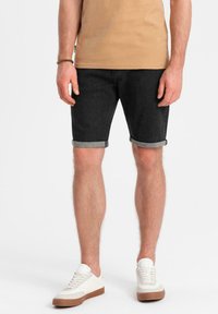 Pantalones cortos de denim negro con dobladillos enrollados, combinados con zapatillas blancas. El conjunto incluye una camiseta de color beige claro y presenta un ajuste casual.