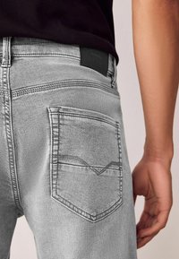 Blugi gri din denim cu un buzunar posterior care are un design distinct cu cusături curbate. O etichetă din piele neagră este atașată în talie.