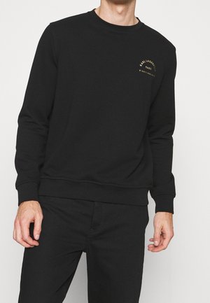Mand iført en sort langærmet sweatshirt med et lille gyldent logo og tekst på venstre bryst, kombineret med sorte bukser.