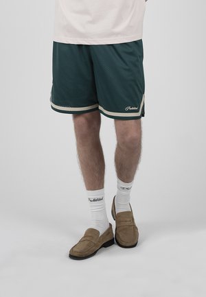 Männliche Beine, die dunkelgrüne Sportshorts, weiße Sneakersocken mit Schriftzug und braune Wildleder-Loafers auf hellgrauem Hintergrund tragen.