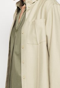 Camicia beige con bottoni e design strutturato, dotata di una tasca sul petto e una blusa verde chiaro sotto, che mette in mostra le trame dei tessuti.