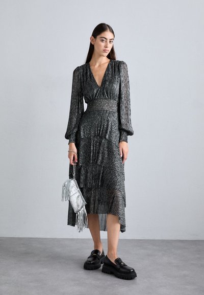 Robe plissée noire métallique à manches longues, décolleté en V profond et jupe à volants. Accessoirisée avec un sac à main argenté à franges et des chaussures noires épaisses.