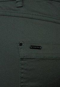 Mörkgrön tyg med en slät textur. Har en sidoficka med en svart metallackent och kontrasterande sömmar längs kanterna.