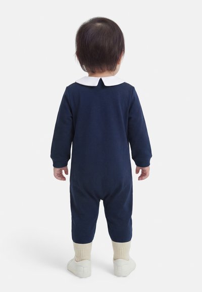 Malenka otroka stoji obrnjena stran, oblečena v mornarskasto modro obleko z belim ovratnikom, kremnimi nogavicami in belimi čevlji na belem ozadju.