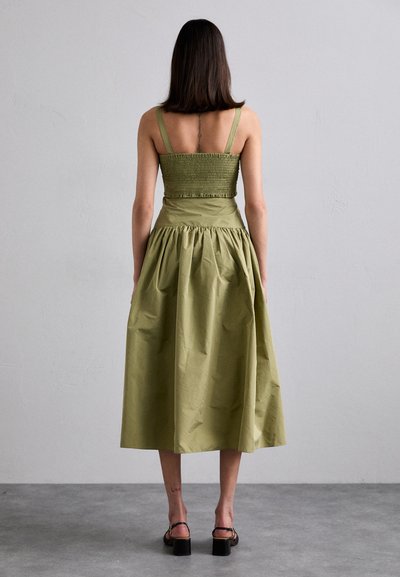 Vestido verde oliva con corsé fruncido, tirantes finos y una falda voluminosa y acampanada que cae por debajo de la rodilla. Sandalias negras de tacón cuadrado.