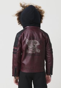 Bambino con capelli ricci che indossa un cappuccio nero e una giacca di pelle bordeaux con una grafica di casco alato e testo sulla schiena, di spalle.