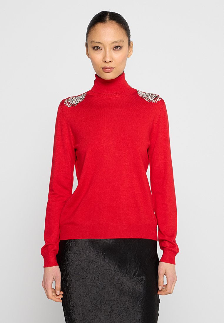 Maglione a collo alto rosso realizzato in tessuto morbido, con spalle decorate e dettagli ornamentali. Indossato con una gonna nera con texture.