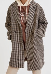 Double-breasted geruite jas in bruine en beige tinten, met een grote reverskraag en zijzakken, gedragen over een bruine hoodie.