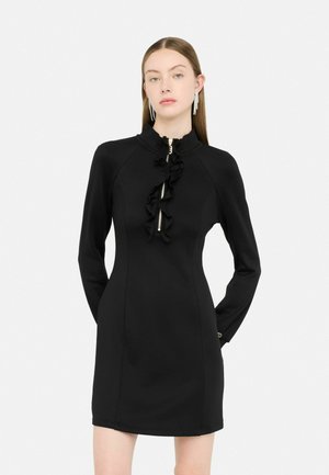 Robe noire à manches longues avec col montant, fermeture éclair à l'avant et détail volanté au centre, confectionnée dans un tissu lisse et extensible.