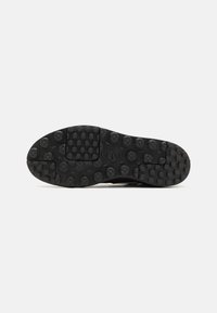 Semelle noire d'une basket Nike montrant des crampons texturés circulaires et carrés pour l'adhérence, vue à plat sur un fond blanc uni.