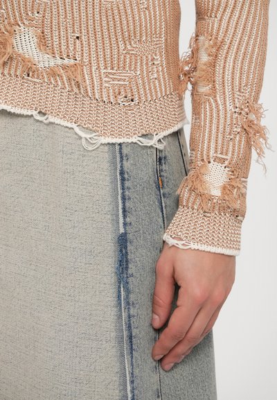 Mână purtând o fustă din denim albastru deschis stă lângă un pulover bej tricotat cu găuri desprinse și margini uzate.