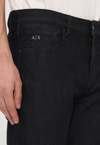 Pantaloni din denim închis, cu o croială slim, având un logo A|X subtil, cinci buzunare și închidere standard cu fermoar metalic și nasture.