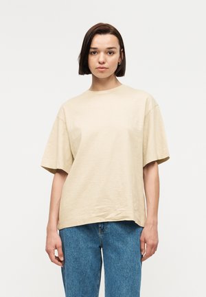 Mlada ženska s kratkimi temnimi lasmi, oblečena v bež preveliko majico in modre kavbojke, stoji ob enostavni svetli podlagi.