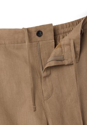 Beige broek met knoopsluiting, gedeeltelijk open ritssluiting, trekkoord in de taille en elastische taillebanddetail op een witte achtergrond.