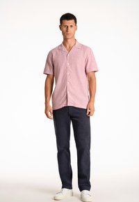 Korte mouwen, lichtroze blouse met een relaxte pasvorm, gecombineerd met donkerblauwe broeken en witte sneakers op een effen achtergrond.