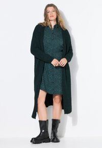 Lange, donkergroene cardigan over een teal gekleed met luipaardprint. Zwarte enkellaarsjes met een chunky zool. Gladde stoftextuur en losse pasvorm.