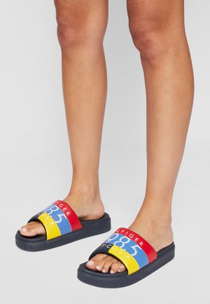 Nogi w wielokolorowych klapkach Tommy Hilfiger z czerwonymi, niebieskimi, żółtymi paskami i czarnymi podeszwami na szarym tle.
