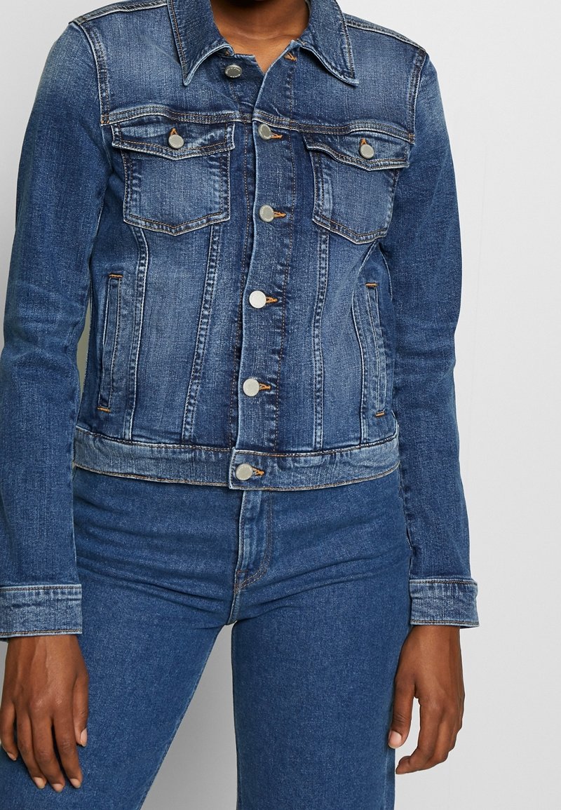 Blå denimjacka med knappstängning, två bröstfickor och sidfickor. Har en klassisk krage och en figurnära design med en något nötta yta.