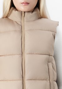 En person med blondt hår iført en beige dunvest med høy krage som er glidd igjen, over en hvit langermet topp, vist fra skuldrene til haken.