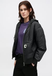 Schwarze Pufferjacke mit Kapuze, ausgestattet mit einem Reißverschluss vorne, gerippten Bündchen und einem Logo-Patch. getragen über einem lila T-Shirt mit grafischem Detail.