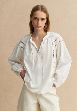 Jonge vrouw met steil blond haar draagt een losse witte blouse met kanten details en crème wijde broekspijpen tegen een beige achtergrond.