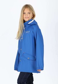 Enfant blonde portant une veste de pluie bleue Swedemount avec capuche et poches avant, regardant l'appareil photo, sur fond blanc.
