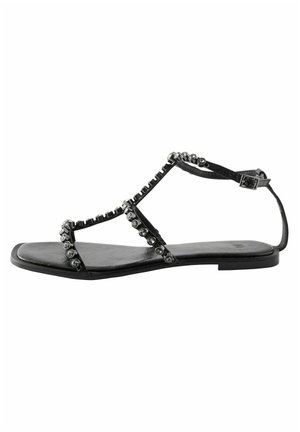 Zwarte platte sandaal met T-strap ontwerp, voorzien van decoratieve metalen studs langs de banden en een glad, textuurvoetbed.