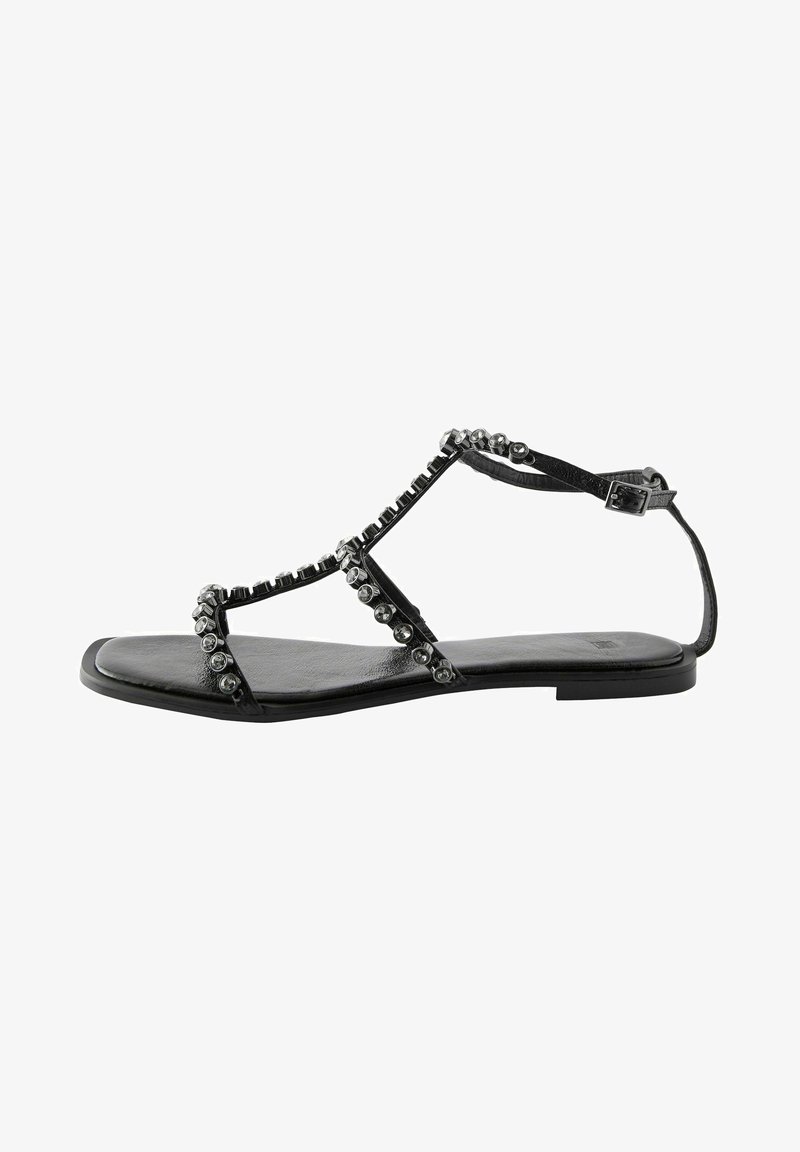 Sandale plate noire avec un design T-strap, ornée de clous métalliques décoratifs le long des lanières et d'une semelle lisse et texturée.