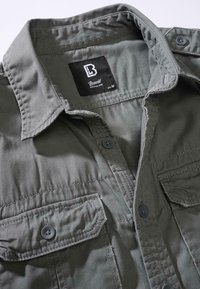 Camicia grigia scura con bottoni, dotata di due tasche sul petto, colletto e etichetta nera di dimensioni medie. Tessuto morbido e strutturato con cuciture sottili.