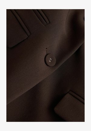 Material maro închis cu un singur nasture rotund și două buzunare drepte, evidențiind o textură netedă și detalii de cusături croite.