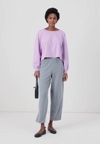 Sweatshirt lung cu maneci lungi de culoare lila, scurt, asortat cu pantaloni largi gri. Ținuta prezintă o textură moale și o croială lejeră. Inclusă este o geantă de umăr neagră.