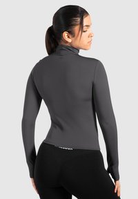 Kvinde med mørkt hår i hestehale iført en tætsiddende grå langærmet top og sorte leggings, vendt lidt væk mod en ensfarvet baggrund.