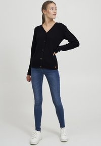 Námořnický pletený kardigan s výstřihem do V, pěti hnědými knoflíky a žebrovanými manžetami, kombinovaný s modrými skinny džínami a bílými teniskami.