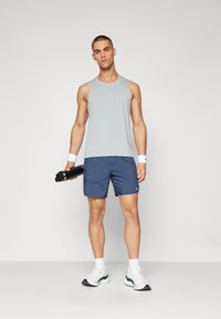 Hellgraues Tanktop und navyfarbene Shorts in athletischer Passform, gefertigt aus atmungsaktivem Material. Weiße Sneakers mit grünen Akzenten. Hält eine schwarze Wasserflasche.