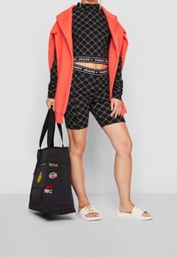 Svart mönstrad träningskläder-set med korallfärgad jacka, vita tofflor och en svart tote-väska med logotyper och lappar.