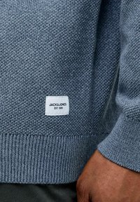 Grå strikket genser med ribbestrikkede mansjetter og kant. Har en hvit etikett med "JACK&JONES" og "EST. 1990." Nærbilde av teksturert stoff.