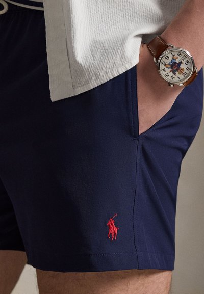 Άτομο που φοράει σκούρο μπλε σορτς με κόκκινο λογότυπο Polo Rider και ρολόι με καφέ δερμάτινο λουράκι και απεικόνιση αρκούδας στο καντράν.