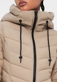 Padded jacket v béžové barvě s vysokým límcem, prošívaným vzorem, černým zipem a stahovacími šňůrkami s ozdobnými konci na kapuci.