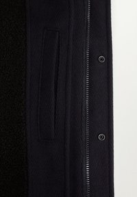 Partie d'un manteau en laine noire avec poche verticale, fermeture éclair métallique et boutons-pression sur un fond uni clair.
