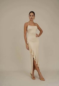 Vestido de cetim creme com uma silhueta ajustada, alças finas, detalhe de ruffle assimétrico na bainha, combinado com sandálias douradas de tiras.