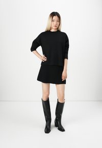 Maglione nero a maglia con spalle scese, abbinato a una mini gonna nera. La modella indossa stivali neri con tacco al ginocchio. Sfondo minimalista.
