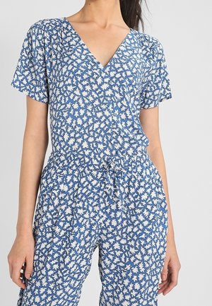 Frau in einem kurzärmeligen Overall mit blau-weißem Blumenmuster, V-Ausschnitt und Kordelzug in der Taille, die vor einem einfarbigen Hintergrund steht.